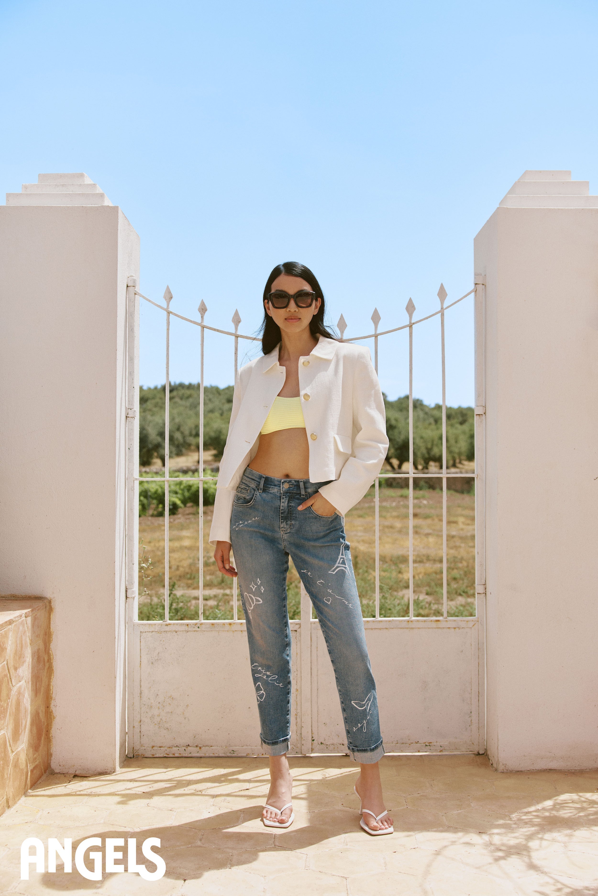 Eine Frau steht entspannt und sommerlich gekleidet da. Sie trägt ein schlichtes Top und darüber eine locker geöffnete Bluse. Dazu kombiniert sie eine lässig sitzende Jeans.